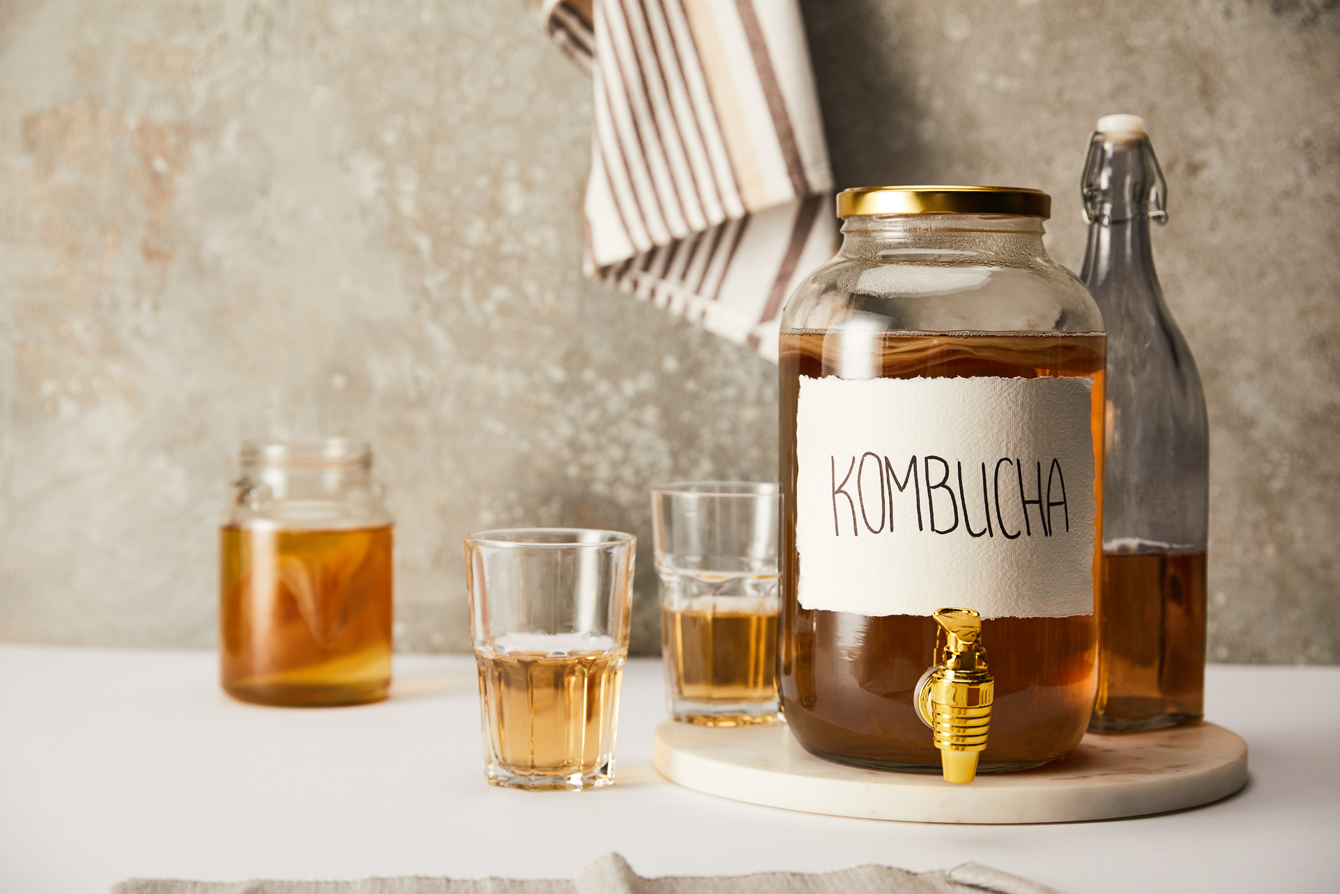 jar with kombucha near glasses and bottle on textured grey background with striped napkin